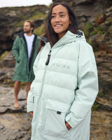 Woman wearing a Vivida Lifestyle All Weather Puffer Changing Robe, Matcha Green Drying Robe for swimming standing in snow. Sticker: Best Premium Changing Robe - The Independent.