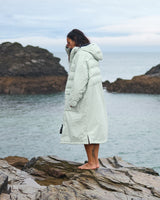 Woman wearing a Vivida Lifestyle All Weather Puffer Changing Robe, Matcha Green Drying Robe for swimming sitting on a beach. Sticker: Best Premium Changing Robe - The Independent.