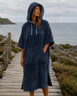 Woman wearing deep navy blue poncho towel in front of the sea, standing on a boardwalk