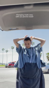 Woman changes clothes wearing a Vivida Original Poncho Towel Changing Robe to change at her car - Mineral Blue / Anthracite Grey. Sticker reads Women's Health's Best Towelling Changing Robe