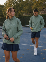 A man and a woman on a blue tennis court wearing matching sage green athletic windbreakers. The woman holds a tennis racket, and the man holds a tennis ball.