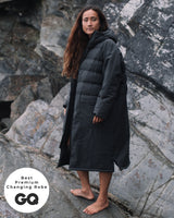 Lead_women - Woman wearing a Vivida Lifestyle All Weather Puffer Changing Robe, Fossil Grey Drying Robe for swimming standing on a rock. Sticker: Best Premium Changing Robe - GQ magazine.