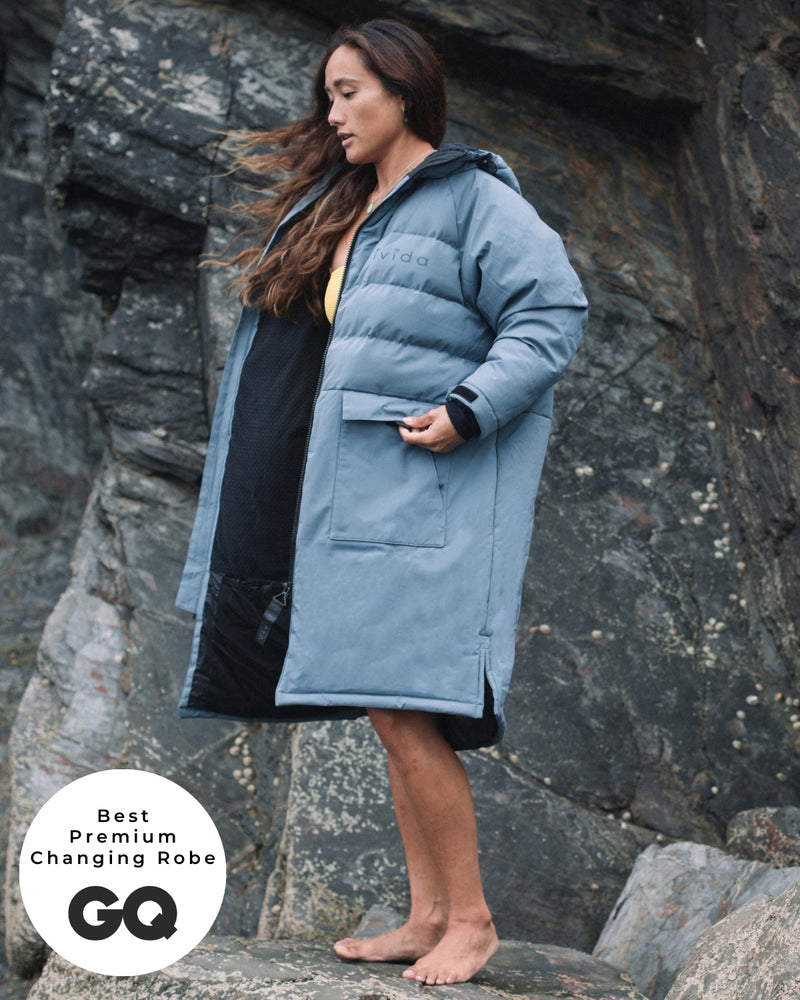 Lead_women - wearing a Vivida Lifestyle All Weather Puffer Changing Robe, Mineral Blue Drying Robe for swimming standing on a rock. Sticker: Best Premium Changing Robe - The Independent and GQ Magazine