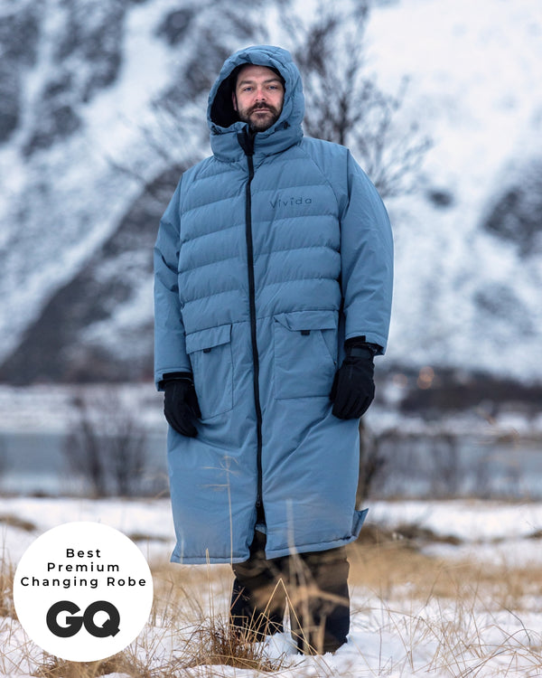 Lead_men - wearing a Vivida Lifestyle All Weather Puffer Changing Robe, Mineral Blue Drying Robe for swimming standing in snow. Sticker: Best Premium Changing Robe - The Independent and GQ Magazine