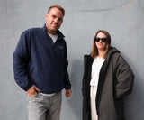 A man and a woman standing side-by-side against a textured gray wall. The man on the left wears a navy blue zip-up jacket with a small white embroidered logo on the chest, paired with light gray jeans.