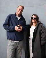 A man and a woman standing against a textured gray wall. The man on the left wears a navy blue zip-up jacket with a small white embroidered logo on the chest, light gray jeans, and a white t-shirt.