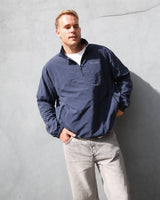 A man wearing a navy blue Vvida quarter-zip pullover jacket and light gray jeans, standing against a textured gray concrete wall.