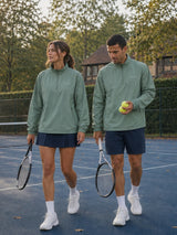 A man and a woman walking on a blue tennis court wearing matching sage green quarter-zip athletic pullovers, navy tennis skirts and shorts, white socks, and white sneakers, with trees in the background.