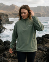 Lead_women. Woman wearing the Vivida Anywhere Lightweight Half-Zip Jacket in Mineral Green on a rocky coast. 