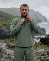 Lead_men. A man wearing an olive green quarter-zip pullover and matching pants stands outdoors on a rocky path with a coastal mountain and ocean in the background, adjusting the collar of the pullover. Vivida Anywhere Lightweight Half-Zip Jacket in mineral green worn on a rainy coastal cliff.